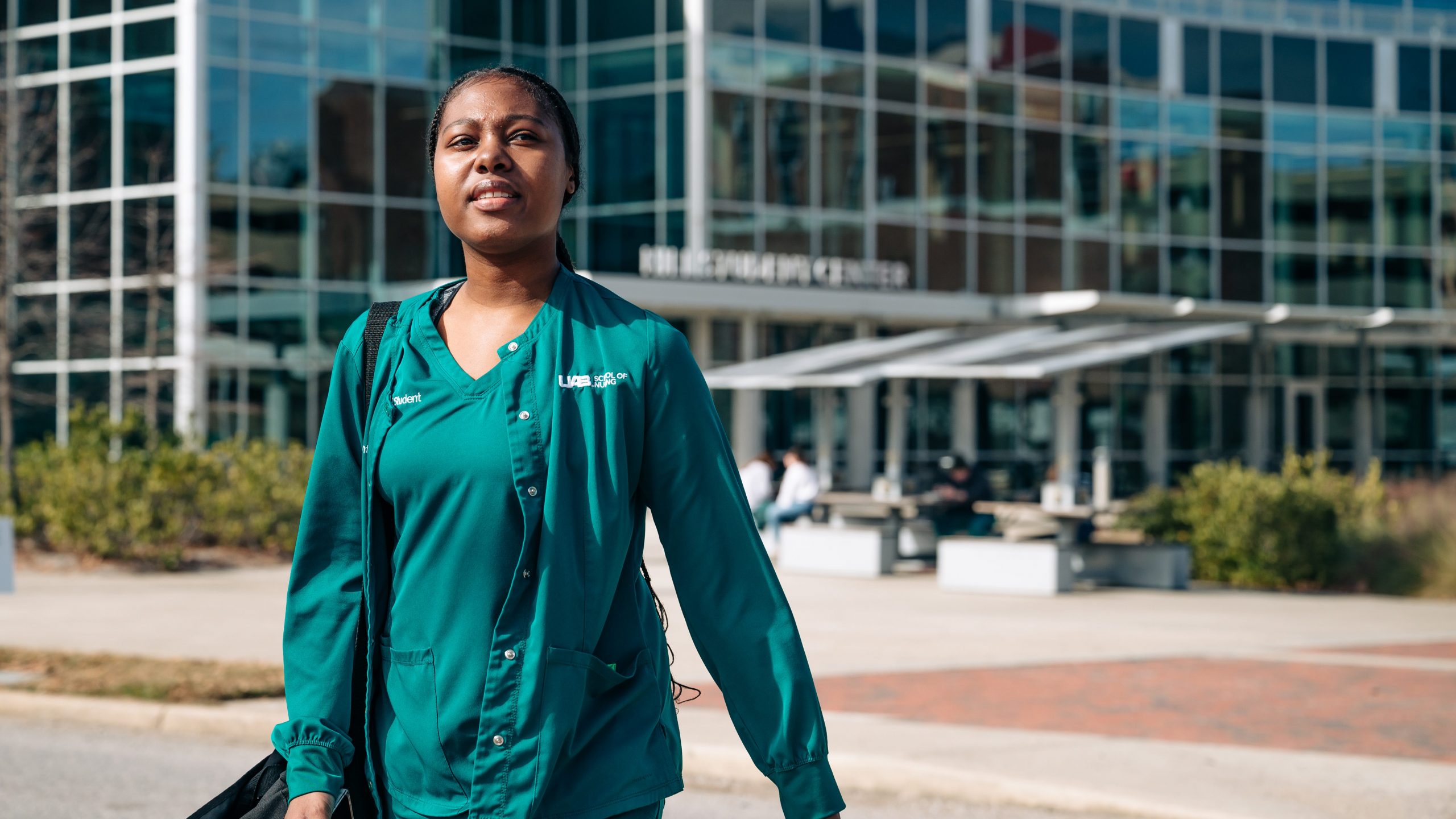 Spring 2025 First Week of Classes. A nursing student is wearing green scrubs while walking on a crosswalk with the Hill Student Center blurred in the background, January 2025.